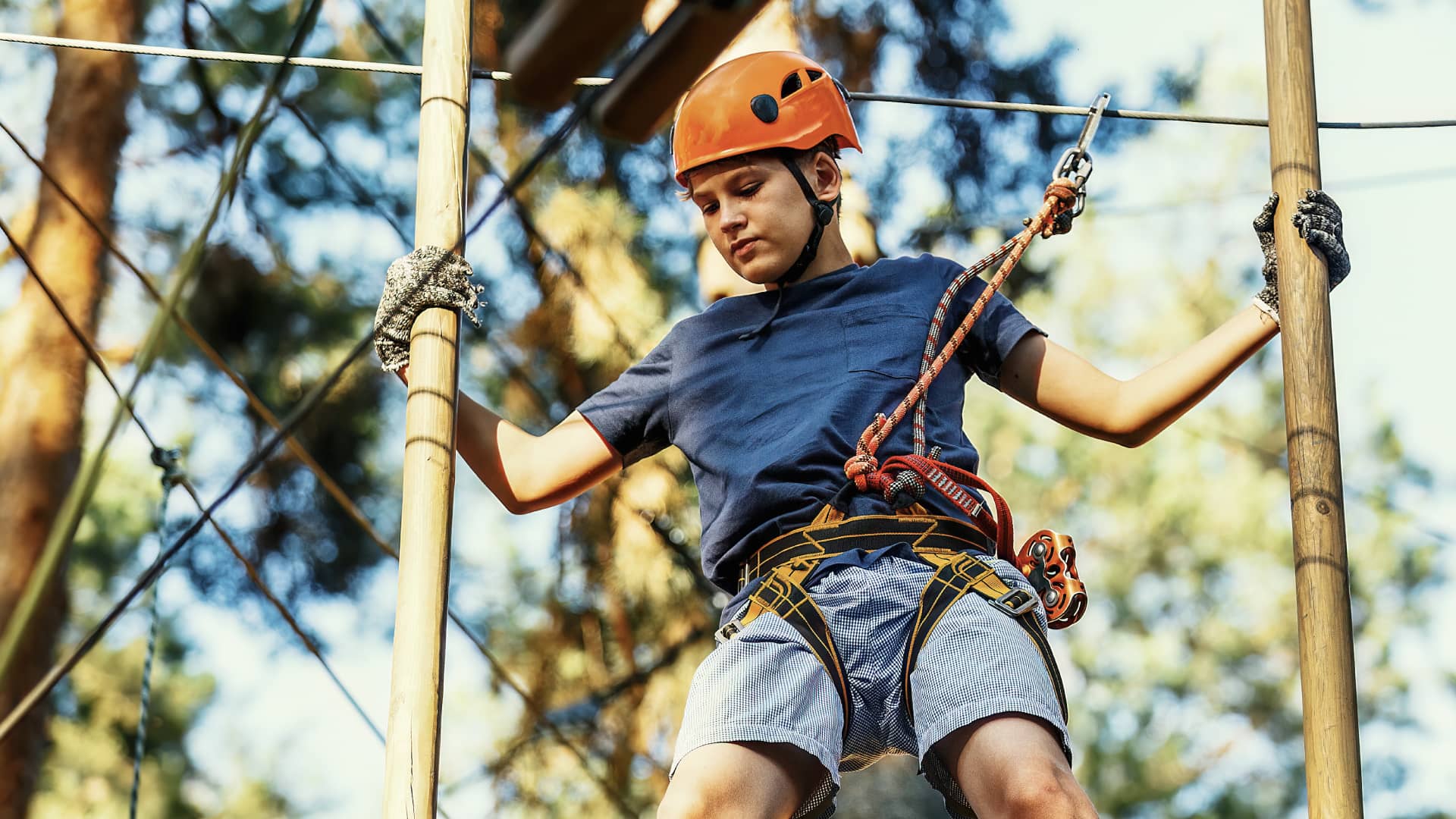 A teenage boy, wearing an orange safety helmet, is engaged in a treetop adventure activity