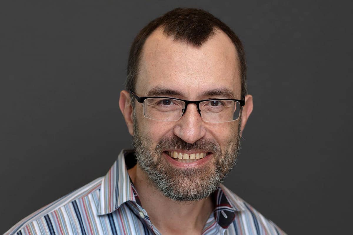 Close-up of a man. The man has short brown hair, and is wearing glasses. He appears to have a well-groomed, graying beard and mustache. He is smiling, and his expression appears friendly. The skin tones are even and natural. His clothing is a light-colored, striped buttoned-down shirt in horizontal stripes of various shades of light beige, gray, and pale blue.